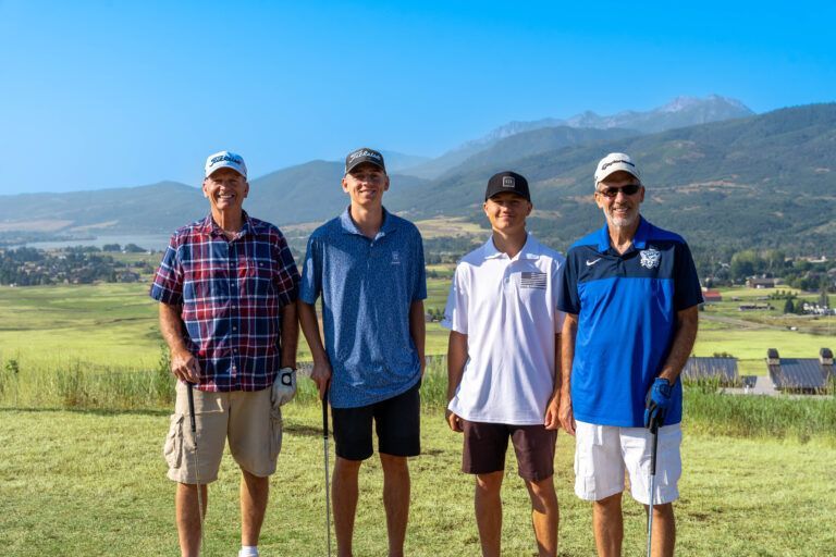 Four people wearing hats and golf attire pose together on a green course with mountains in the background.
