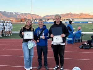 Three people standing on a running track holding certificates, with mountains and a stadium in the background.