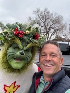 A smiling man takes a selfie next to a person dressed in a Grinch costume wearing a holiday wreath on their head.