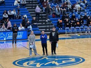 Three people stand on a basketball court with a school logo, in front of blue bleachers filled with spectators.