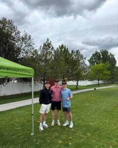 Three individuals stand on a green lawn by a green event tent under a cloudy sky.