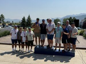 A group of people wearing medals stand on a podium outdoors on a sunny day with mountains in the background.