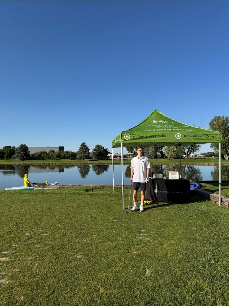 A person stands under a green canopy tent by a lake on a sunny day, with a kayak visible on the water nearby.