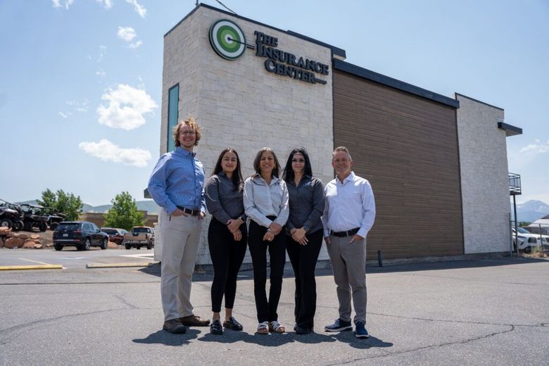 City skyline at sunset with a backdrop of mountains and a foreground of lush, green trees and residential houses.