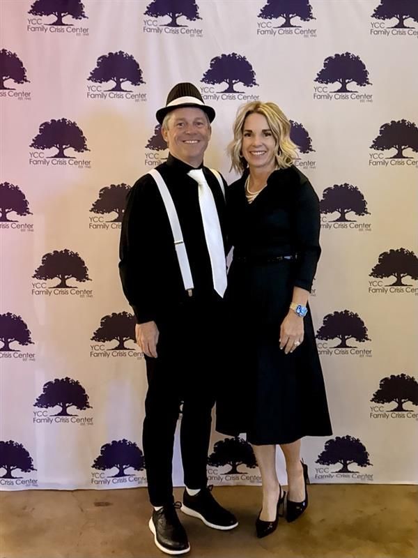 A person in a fedora, white tie, and suspenders stands beside a person in a black dress against a logo-patterned backdrop.