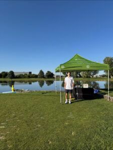 A person stands under a green branded tent at a grassy park beside a lake on a sunny day.