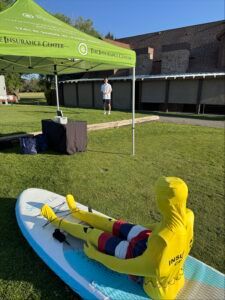 A yellow bodysuit-clad figure sits on a blue paddleboard on a lawn near a green canopy tent and a standing person.
