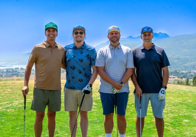 Four men stand together smiling on a sunny golf course with mountains and a lake in the background.