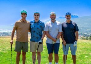 Four people stand side-by-side on a golf course holding clubs, wearing casual apparel against a mountainous landscape.