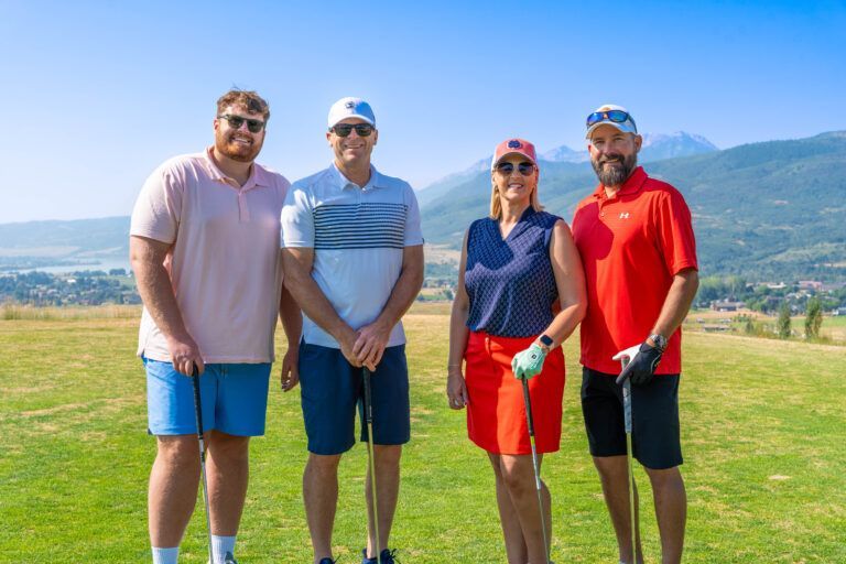 Four people stand in a line on a golf course with mountains in the background, holding golf clubs and smiling.