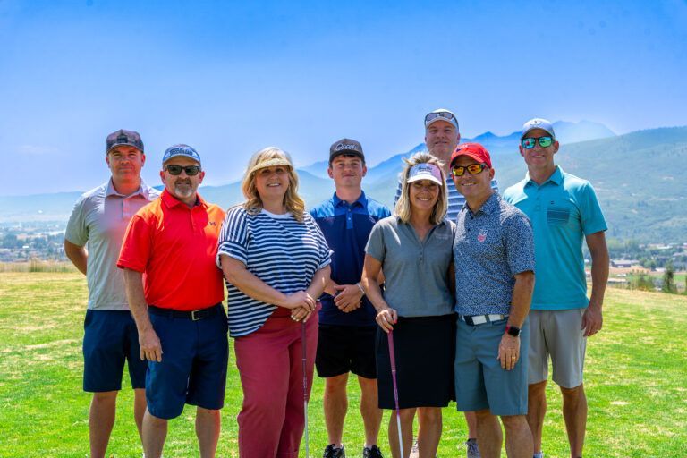 A group of eight people stand in two rows on a sunny golf course, wearing casual attire, with a mountain backdrop.