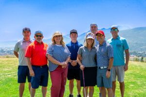 A group of eight people posing together outdoors on a sunny, grassy golf course with mountains in the background.