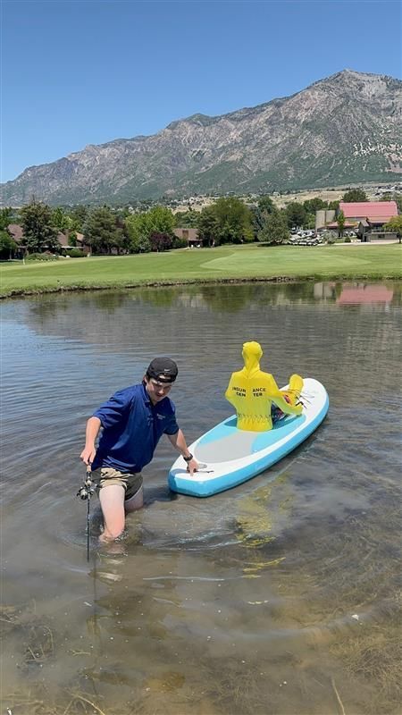 A person in a blue shirt stands in a shallow pond next to a light blue paddleboard holding a yellow mannequin.