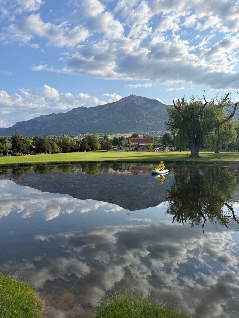 A person in a yellow life vest kayaks on a calm pond reflecting a mountain and cloudy blue sky.