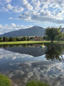 A person paddles a small boat on a still pond, reflecting a large mountain and cloudy blue sky.