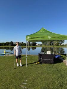 A person stands beside a green tent at a park next to a lake on a sunny day.