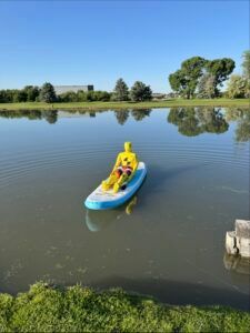 A yellow crash test dummy sits on a light blue paddleboard floating in a calm, reflective pond on a sunny day.