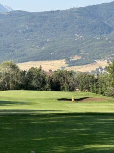 A golf course fairway leading to a green with a bunker and flag, set against a backdrop of mountains and trees.