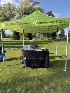 A green branded pop-up canopy tent with a black table setup for The Insurance Center on a grassy golf course.