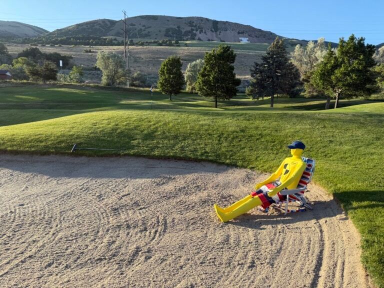 A yellow crash-test dummy wearing a cap relaxes in a beach chair within a sand trap on a sunny golf course.