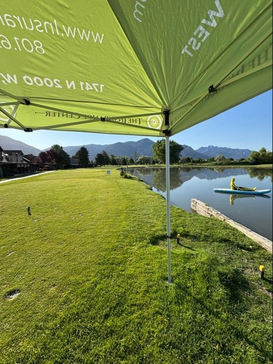 A person paddles on a lake next to a grassy field under the shade of a lime green event tent.