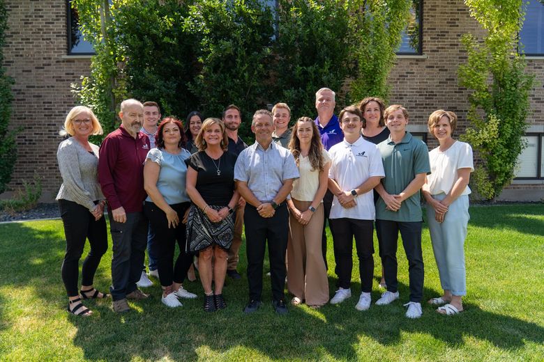Aerial view of a town nestled at the base of a long, red sandstone cliff under a clear blue sky.