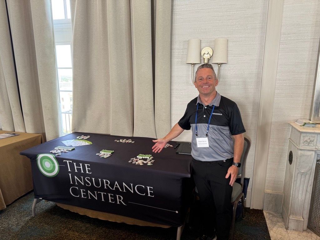 A smiling person in a polo shirt gestures toward a black display table for The Insurance Center in a hotel room.