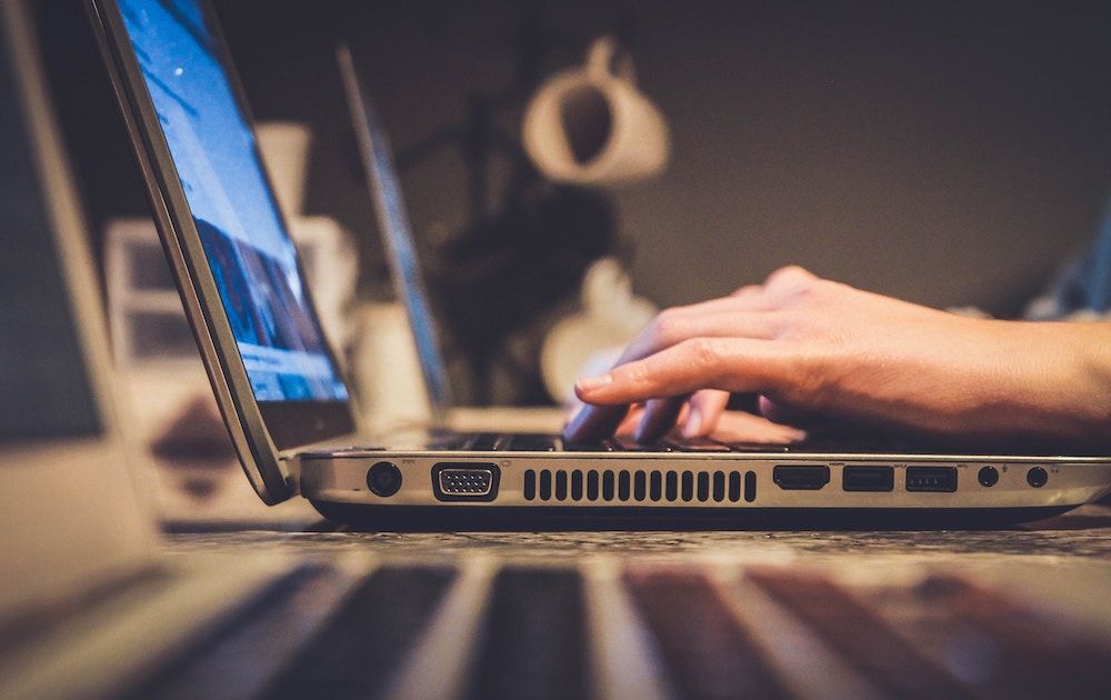 A close-up side view shows a person typing on a laptop, with another laptop screen visible in the soft-focus background.