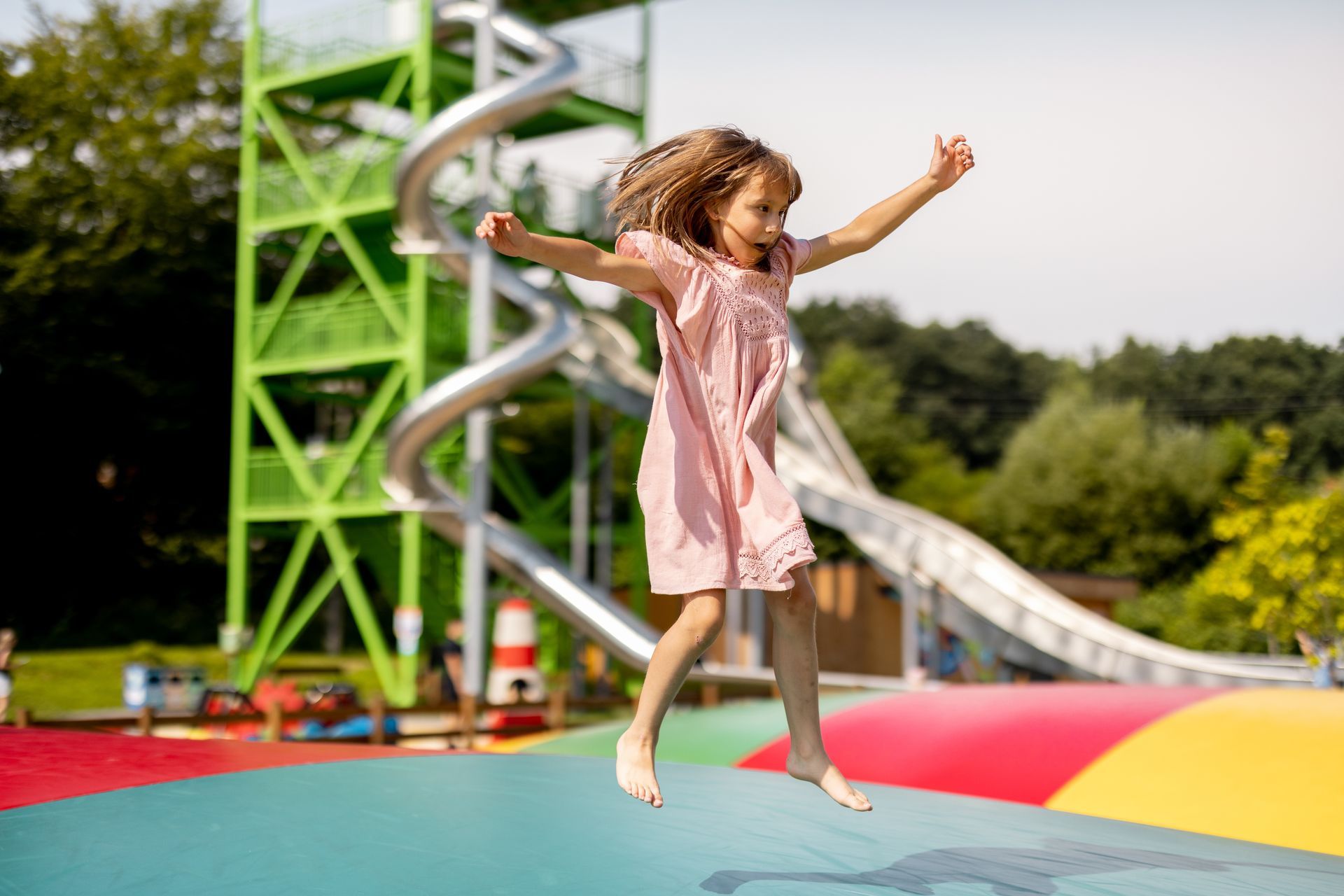 A child in a pink dress jumps high on a colorful inflatable pillow at an outdoor playground with a spiral slide.