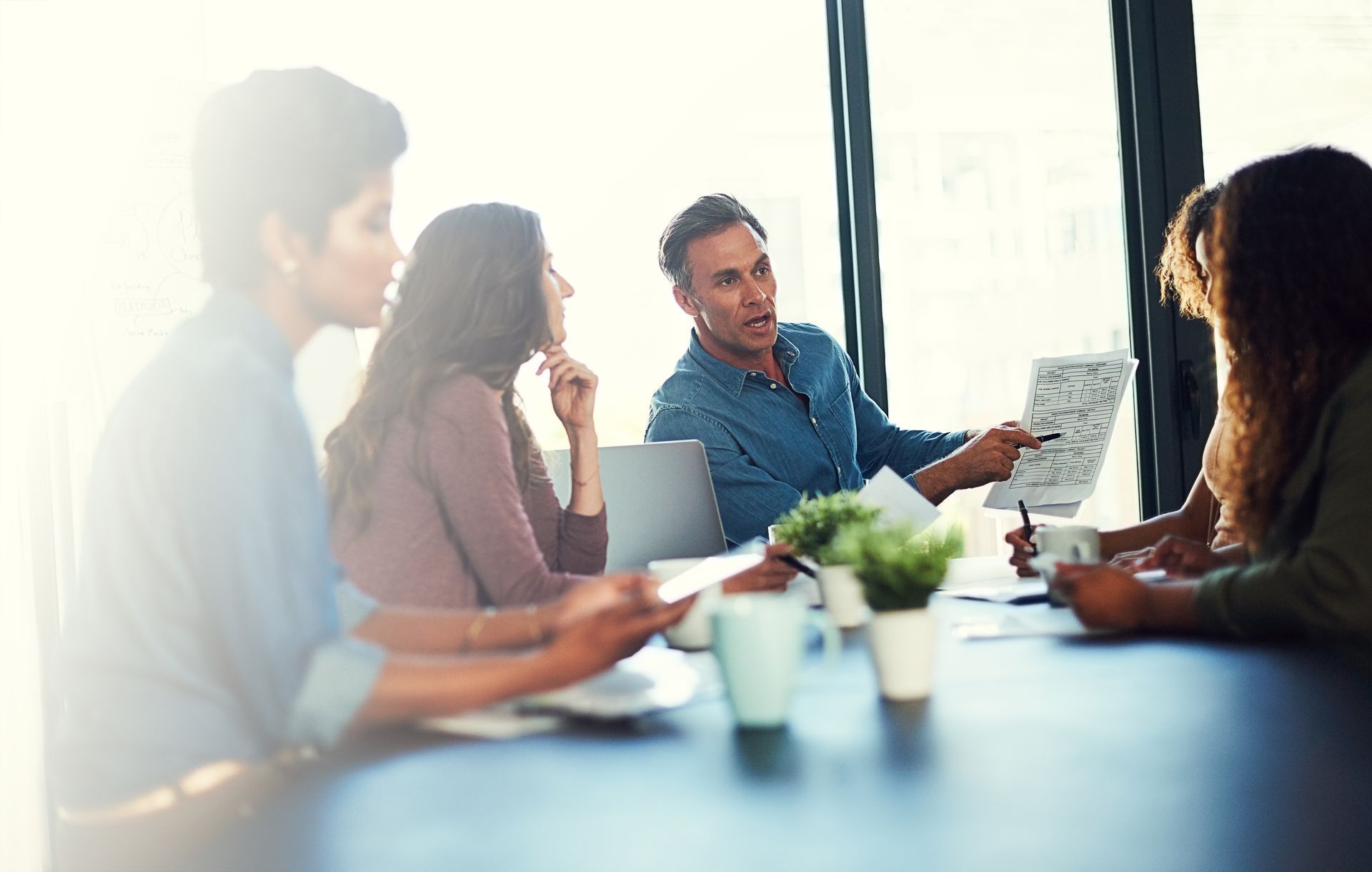 A professional team sits around a conference table in a sunlit office while a colleague points to a document.