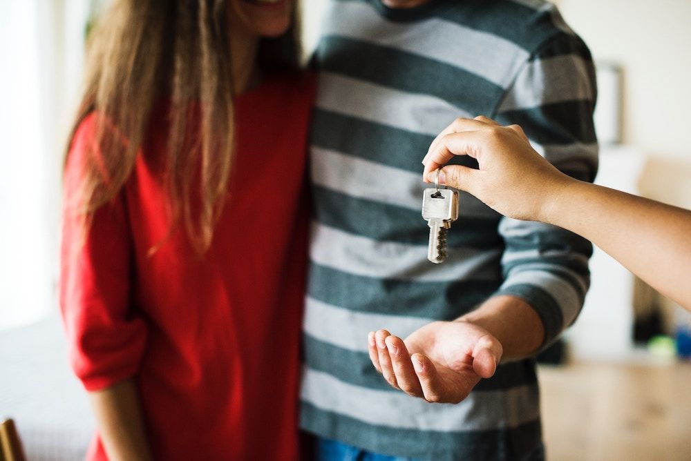 A hand holds out a silver house key toward a couple, who are standing together with their hands open to receive it.