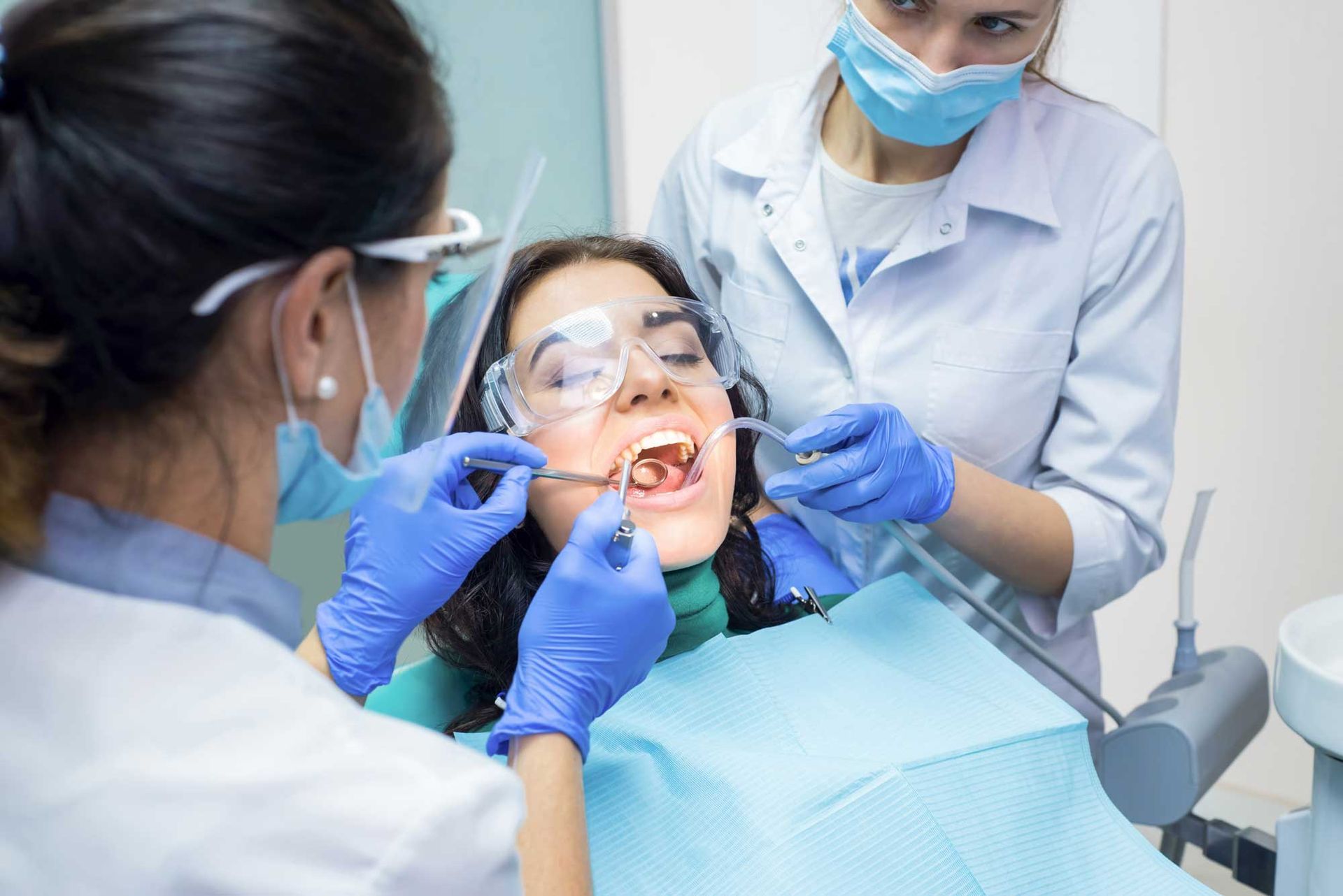 Dentist examining a patient's teeth. Two dental professionals in masks and gloves, patient with mouth open.