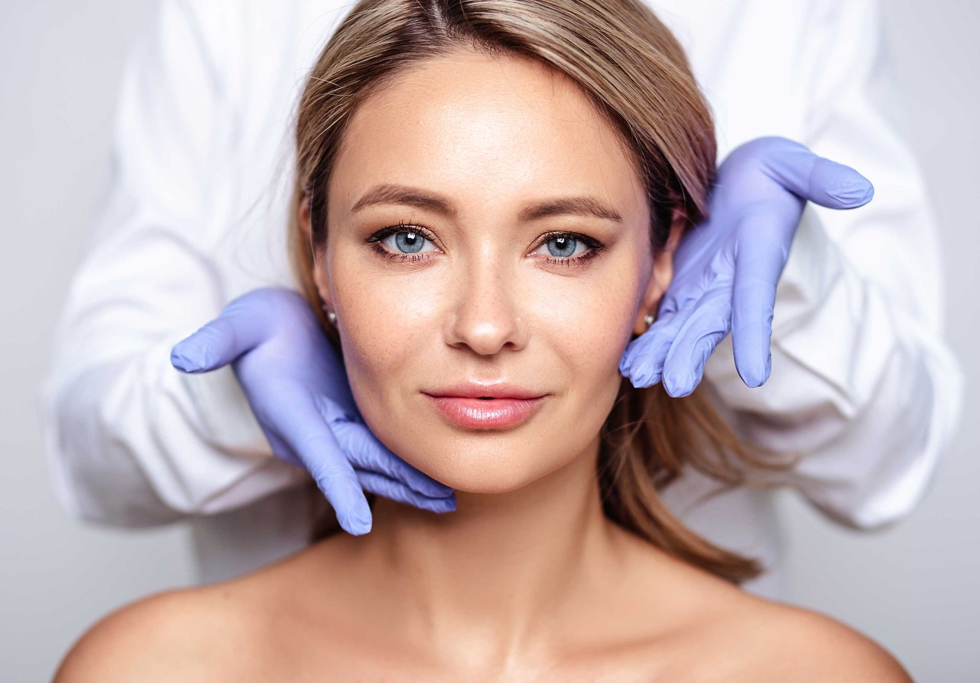 Woman's face examined by someone wearing blue gloves, likely for a cosmetic procedure in a clinic setting.