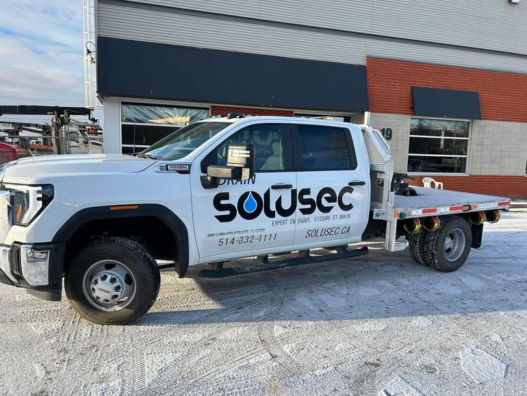 Un camion plateau blanc à cabine double, portant la marque « Solusec », est stationné sur un terrain en gravier devant un bâtiment.
