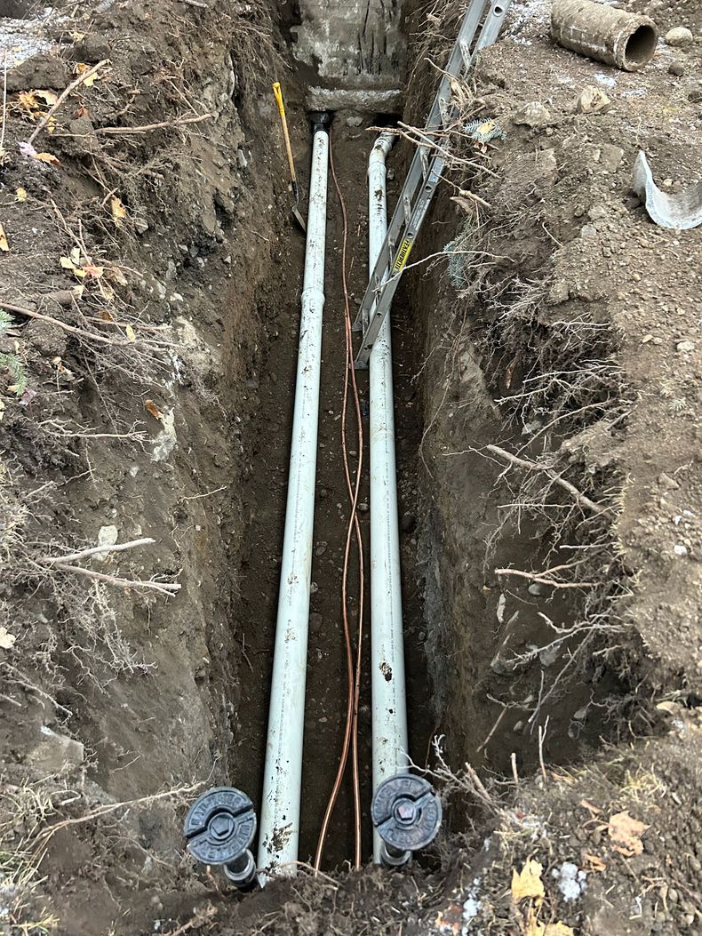 Deux conduits parallèles en PVC gris, aux extrémités bouchées, reposent dans une tranchée profonde et étroite, à côté de fils de cuivre apparents.