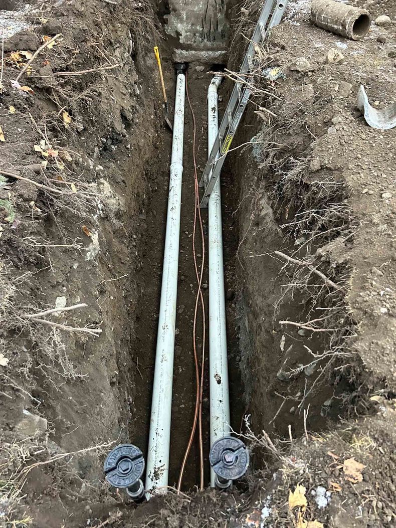 Deux conduits parallèles en PVC gris, aux extrémités bouchées, reposent dans une tranchée profonde et étroite, à côté de fils de cuivre apparents.