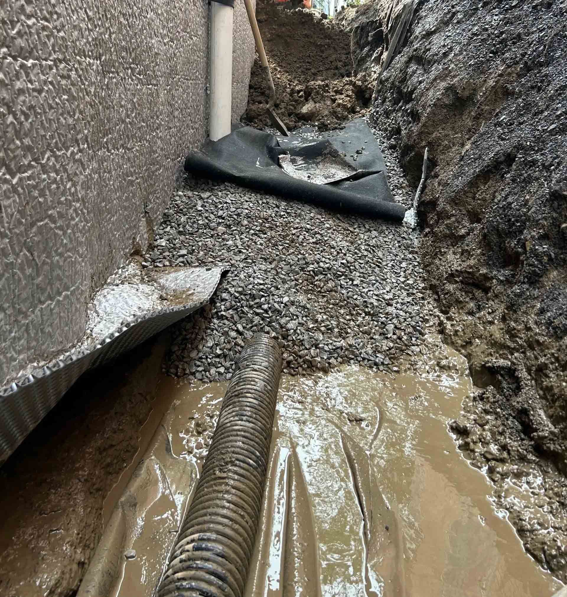 Tranchée étroite et boueuse avec eau stagnante, gravier et tuyaux de drainage noirs le long des parois après le ruissellement.