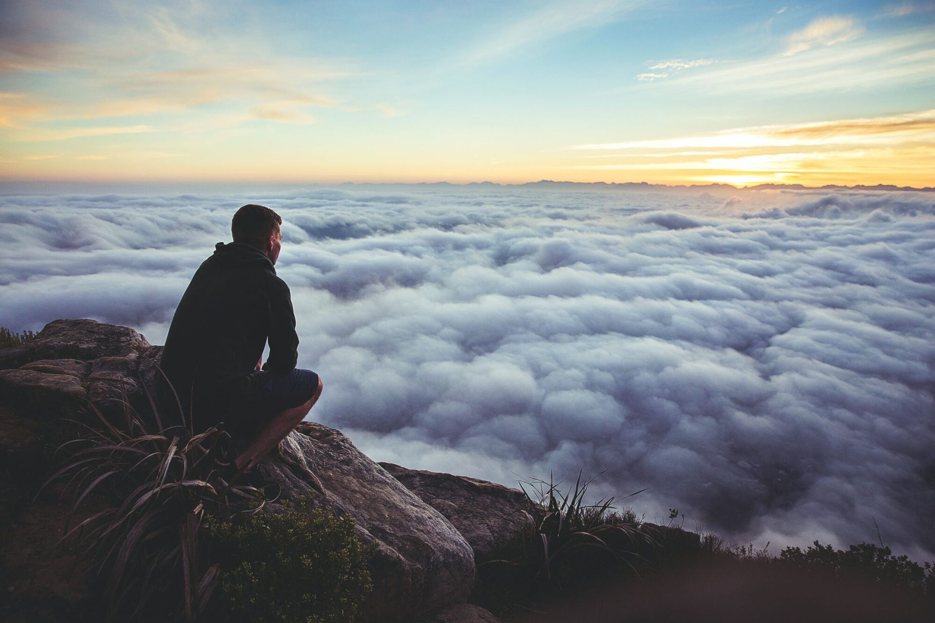 Person seated on a rocky cliff above a sea of clouds at sunrise
