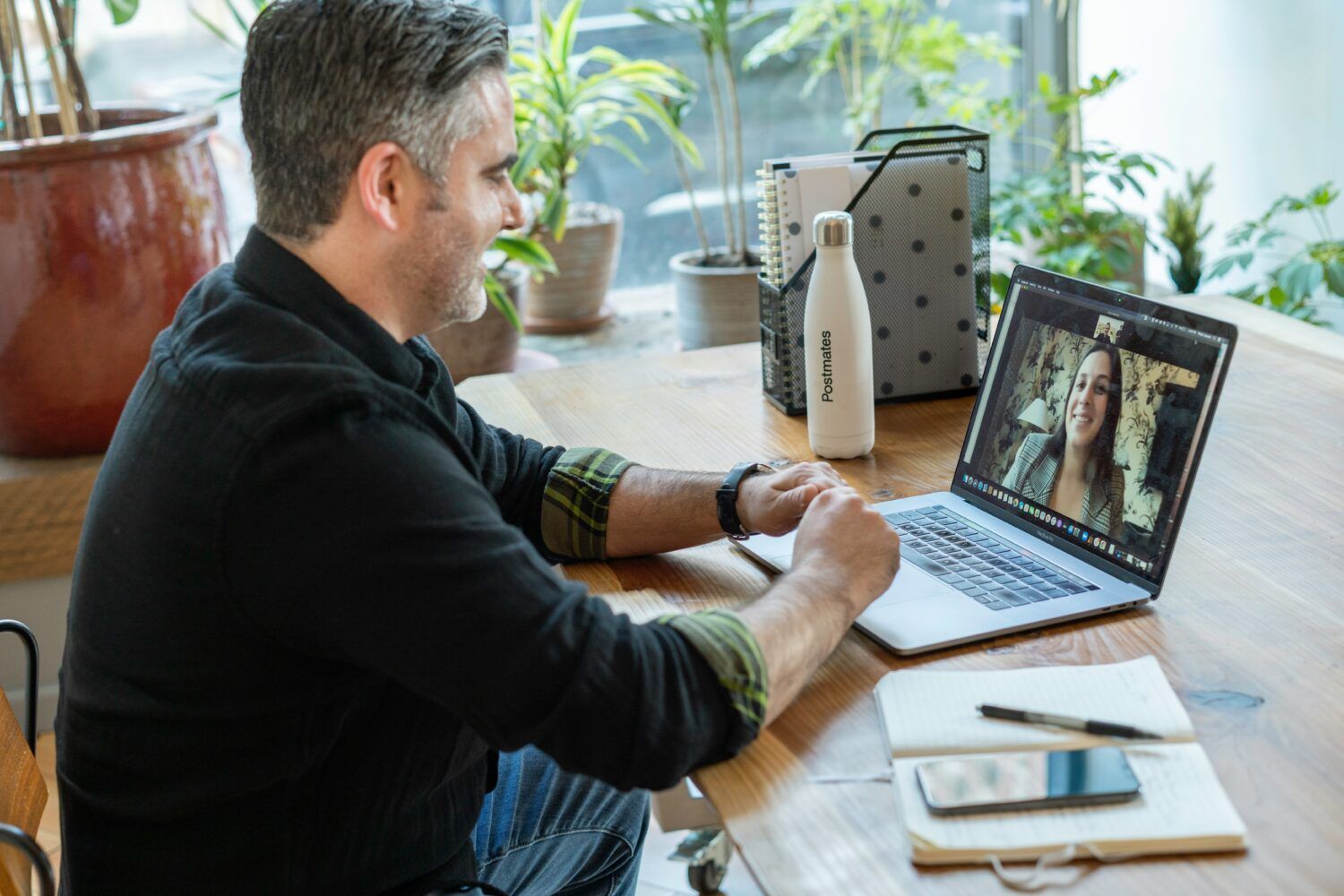 Person working on a laptop at a wooden table in a bright room with plants and a coffee cup.