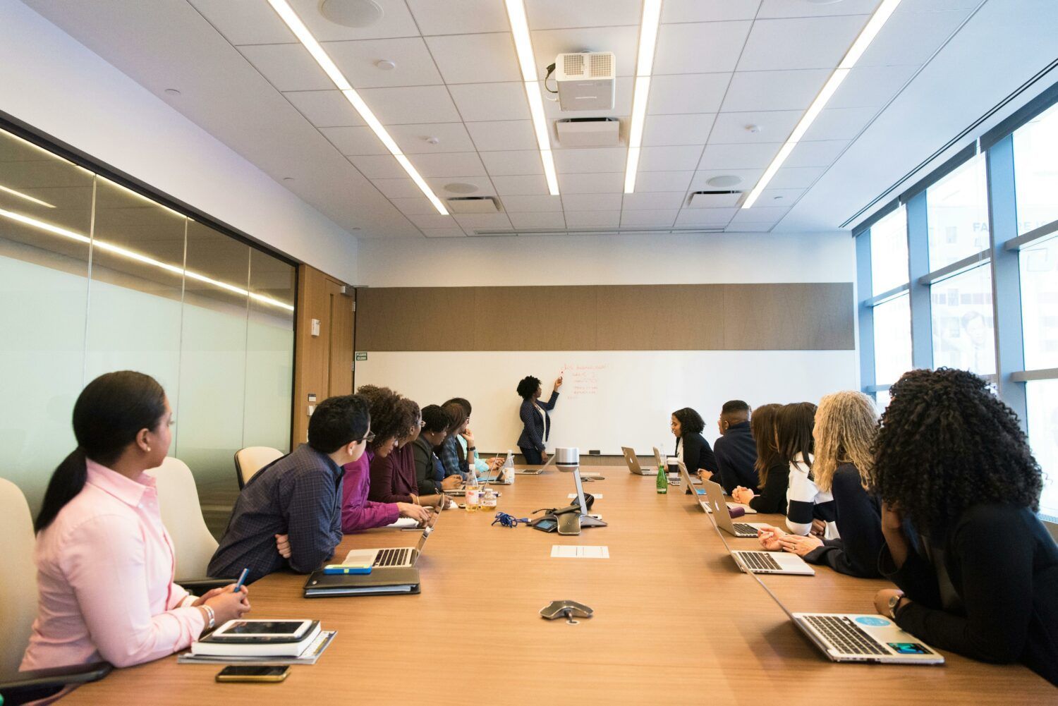 Conference room meeting with people seated around a long table, watching a presenter at the front.