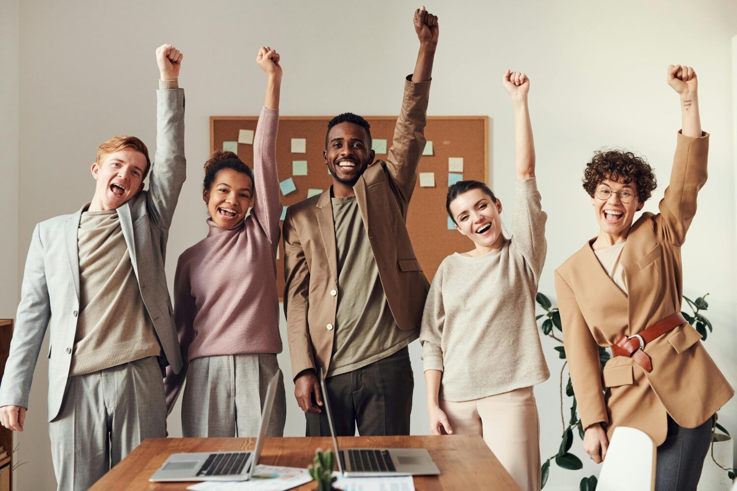 Team in a meeting room celebrating with raised arms around a table and laptop