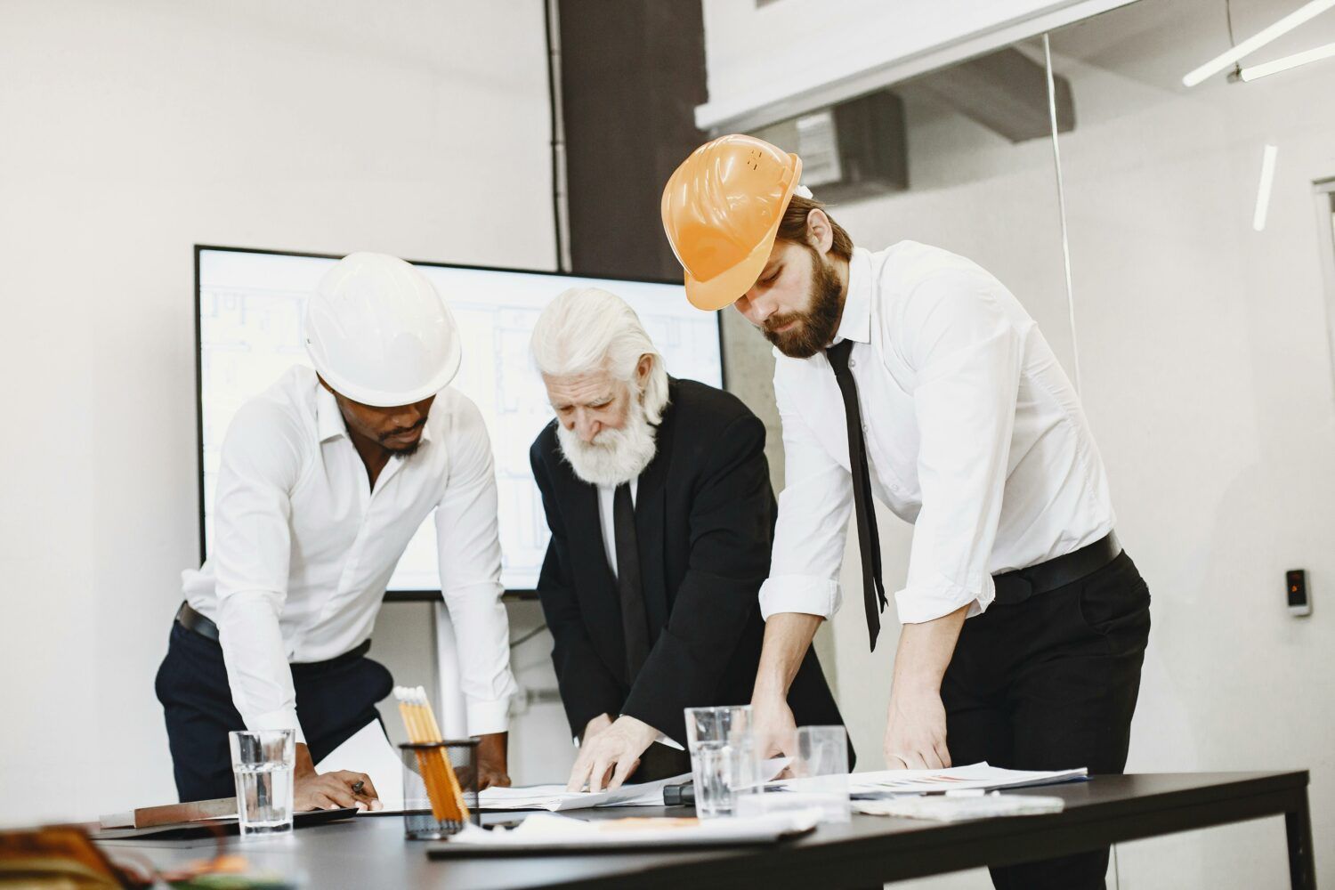 Three coworkers in hard hats reviewing blueprints at a desk in an office under construction
