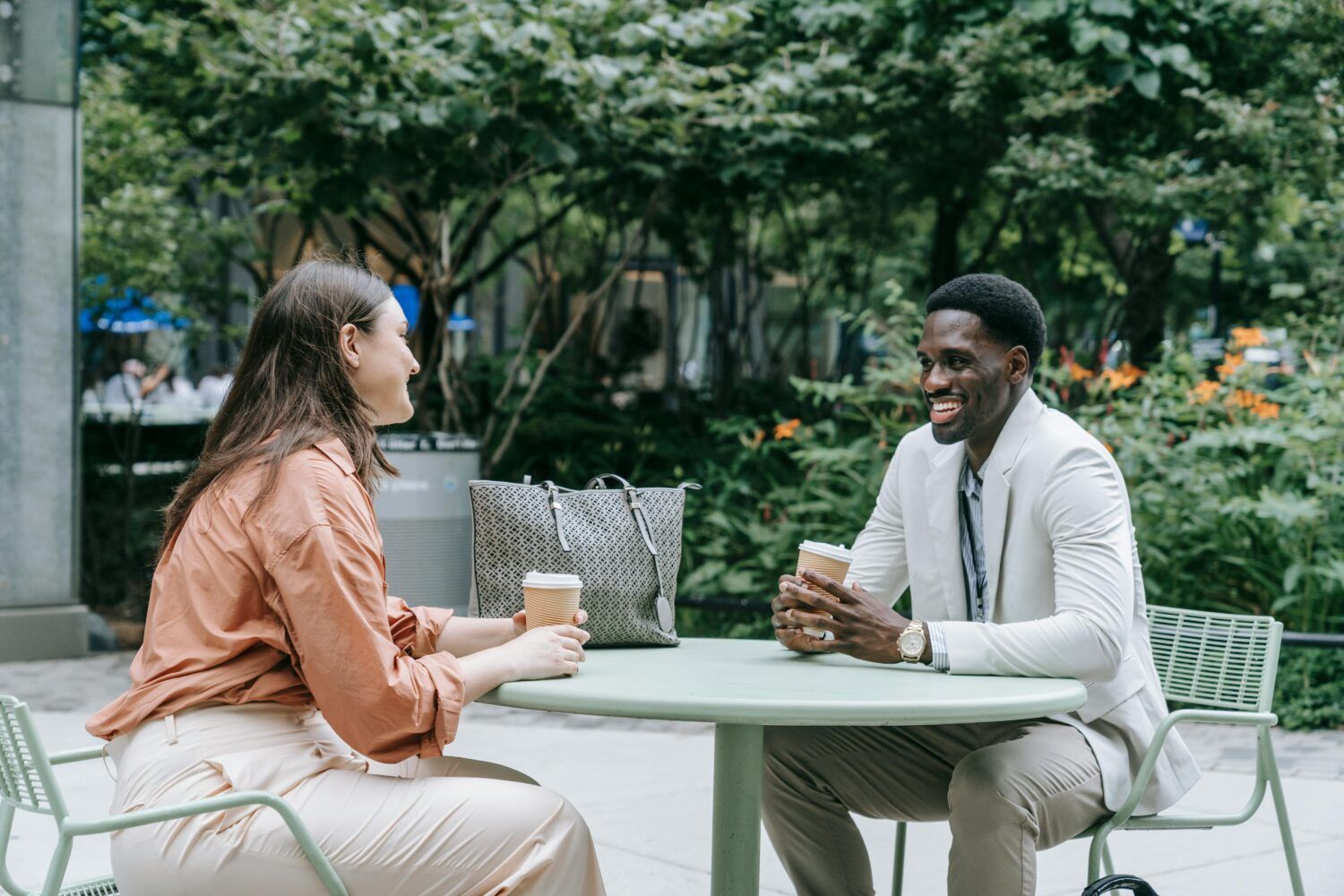 Two people sit at an outdoor table in a green courtyard, smiling and talking over coffee.
