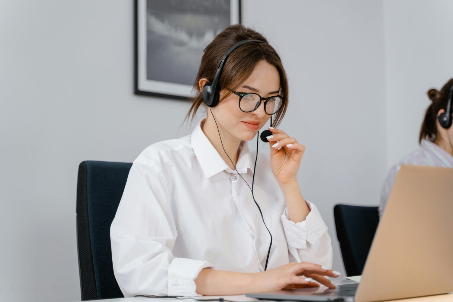 Woman wearing glasses and a headset, working at a laptop in a bright office.