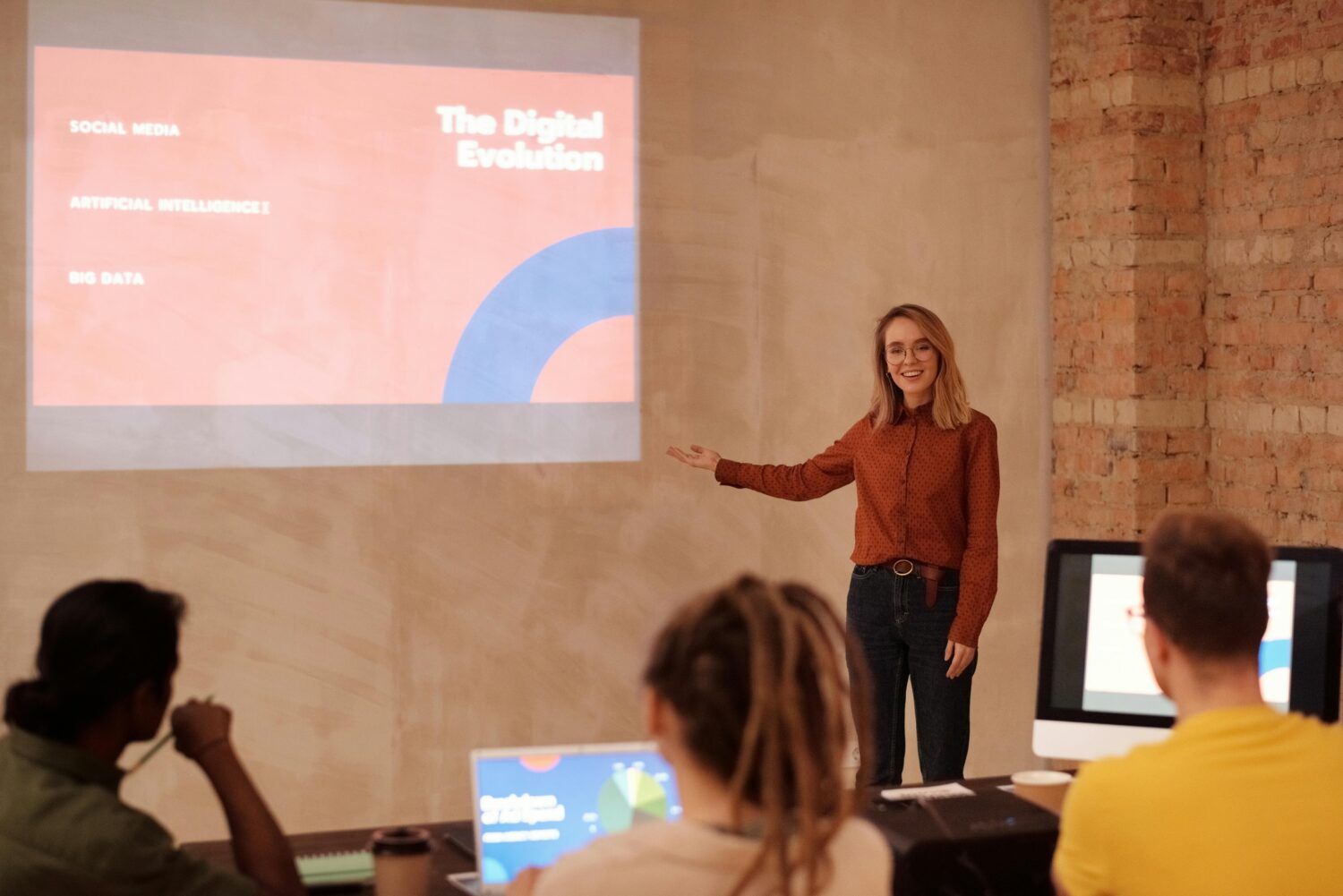 Presenter speaking to seated audience beside a projected slide in a small classroom or meeting room.
