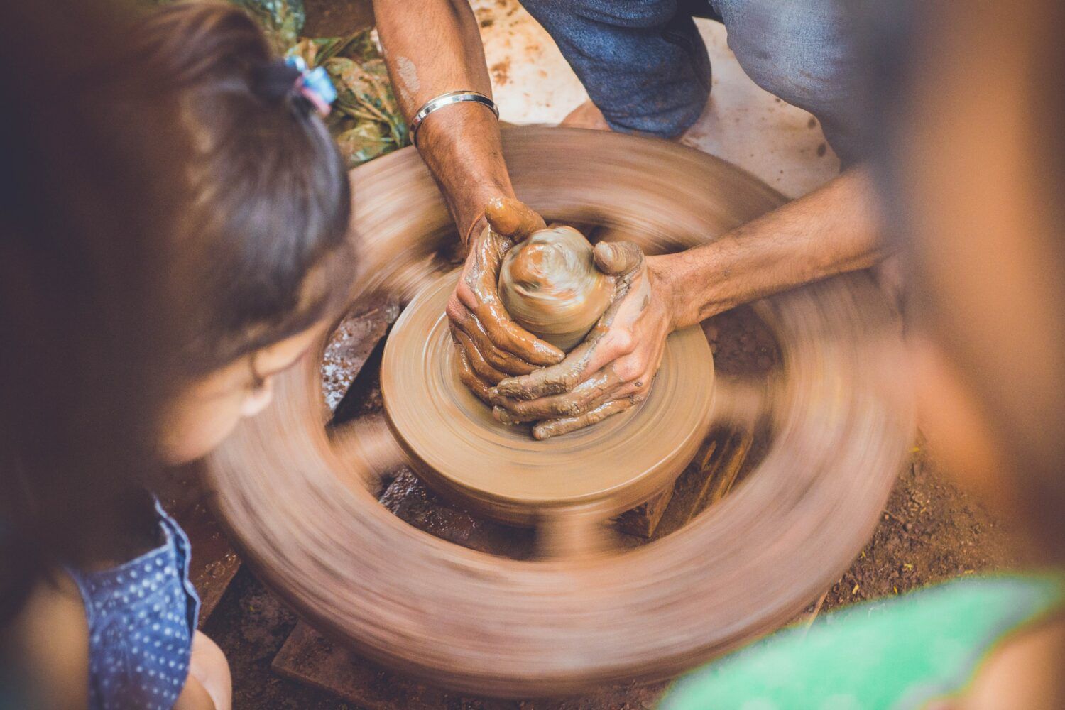 Hands shaping clay on a spinning potter’s wheel, with onlookers nearby.