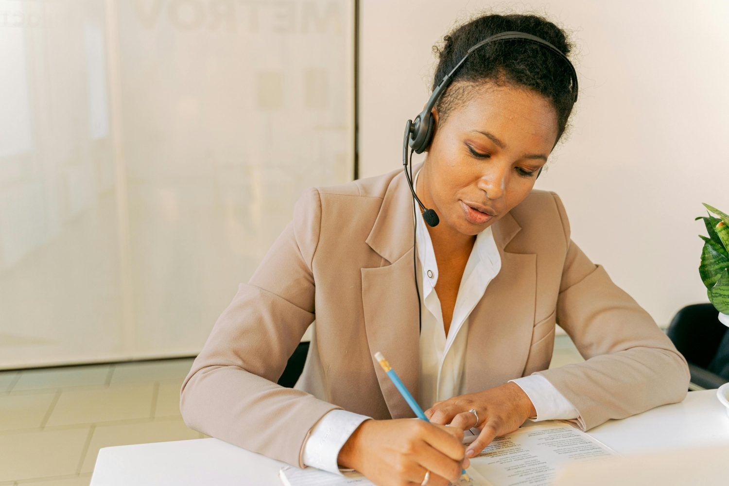Woman in a beige blazer wearing a headset, writing notes at a desk in a bright office.
