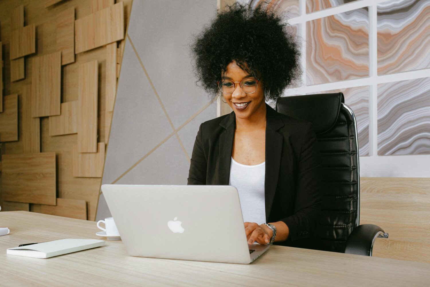 Woman smiling at a desk with a laptop in a modern office, wearing a black blazer.