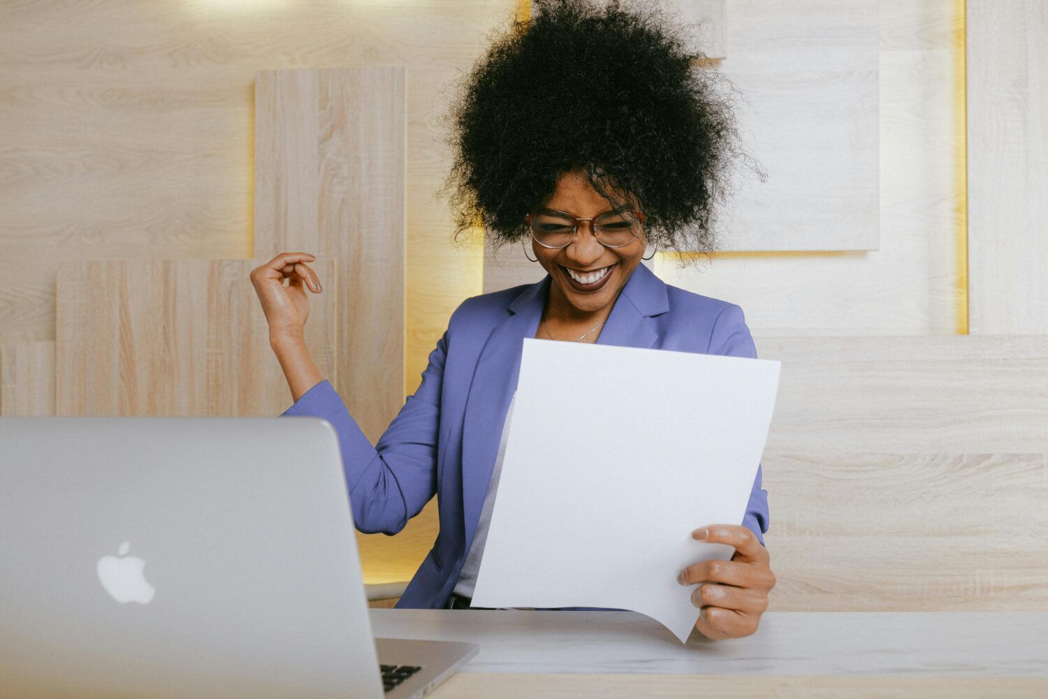 Smiling person in a purple blazer reading papers beside a laptop at a desk