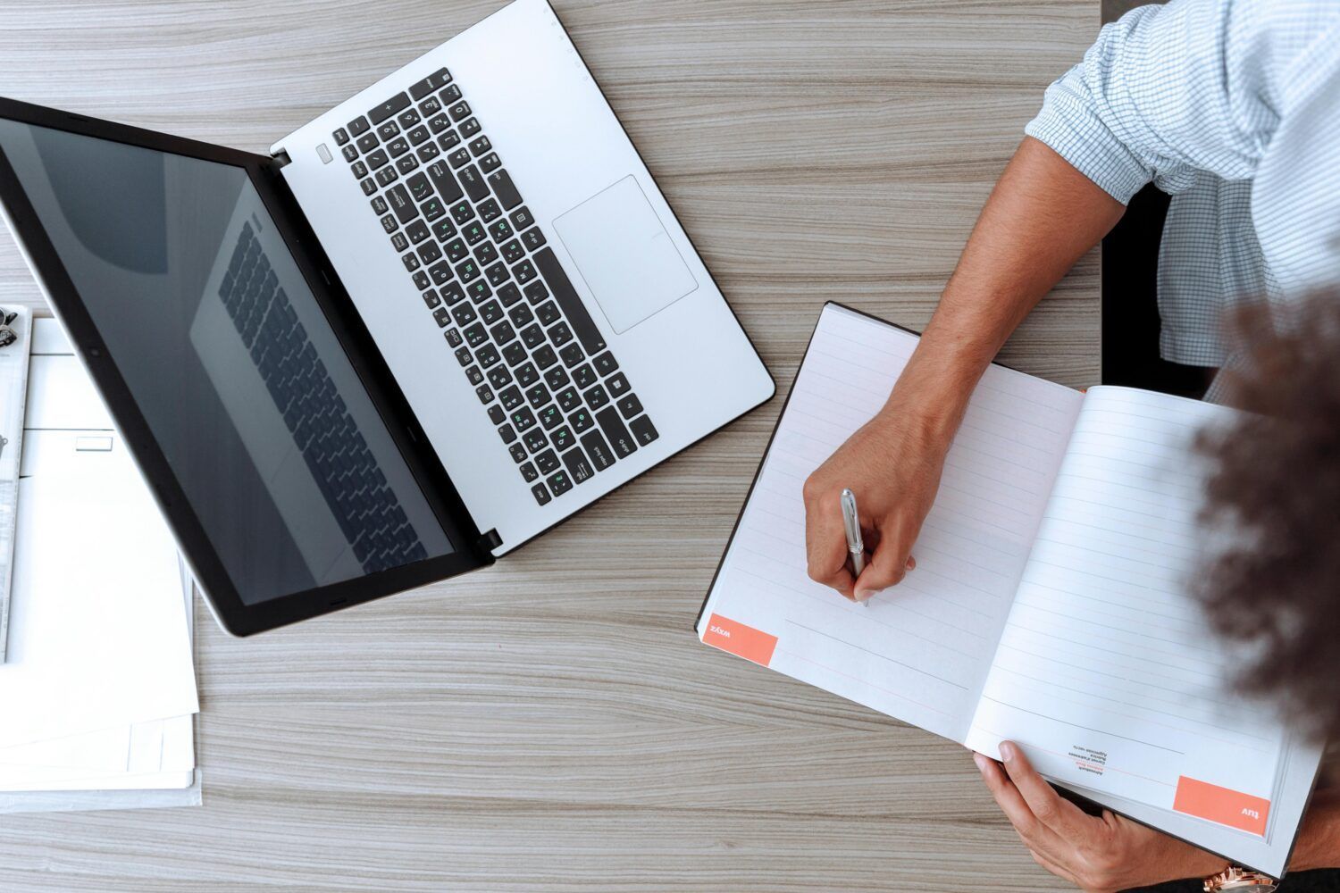 Overhead view of a person writing in a notebook beside a laptop on a wooden desk